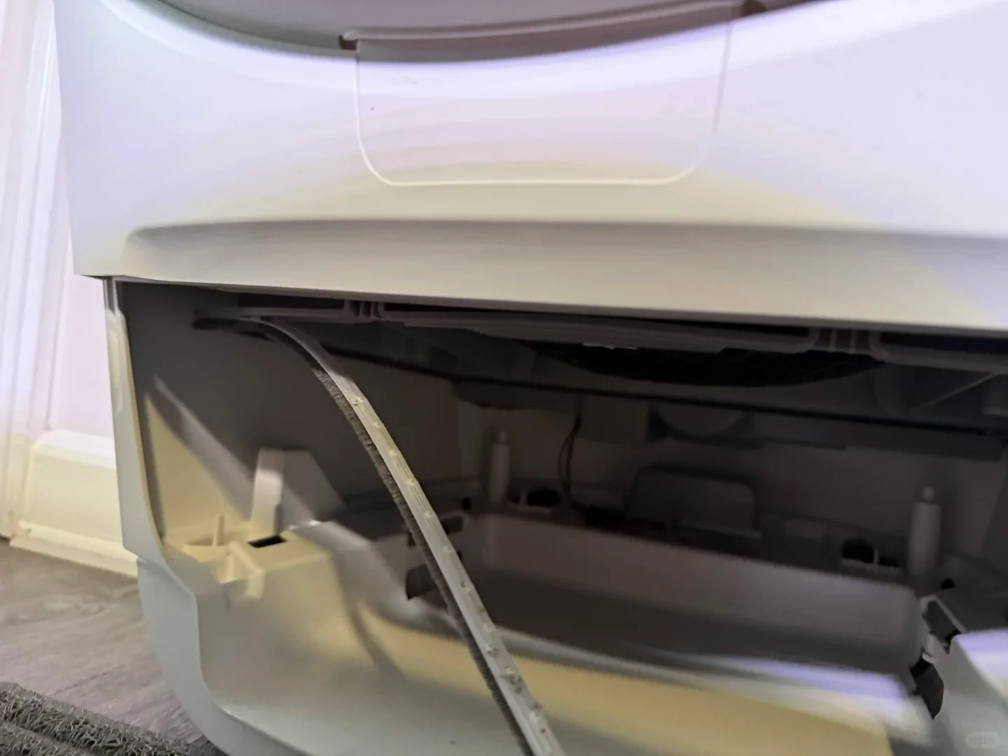 A close-up of the lower interior compartment of a white plastic appliance, where a loose sealing strip or brush seal has detached and is hanging down.
