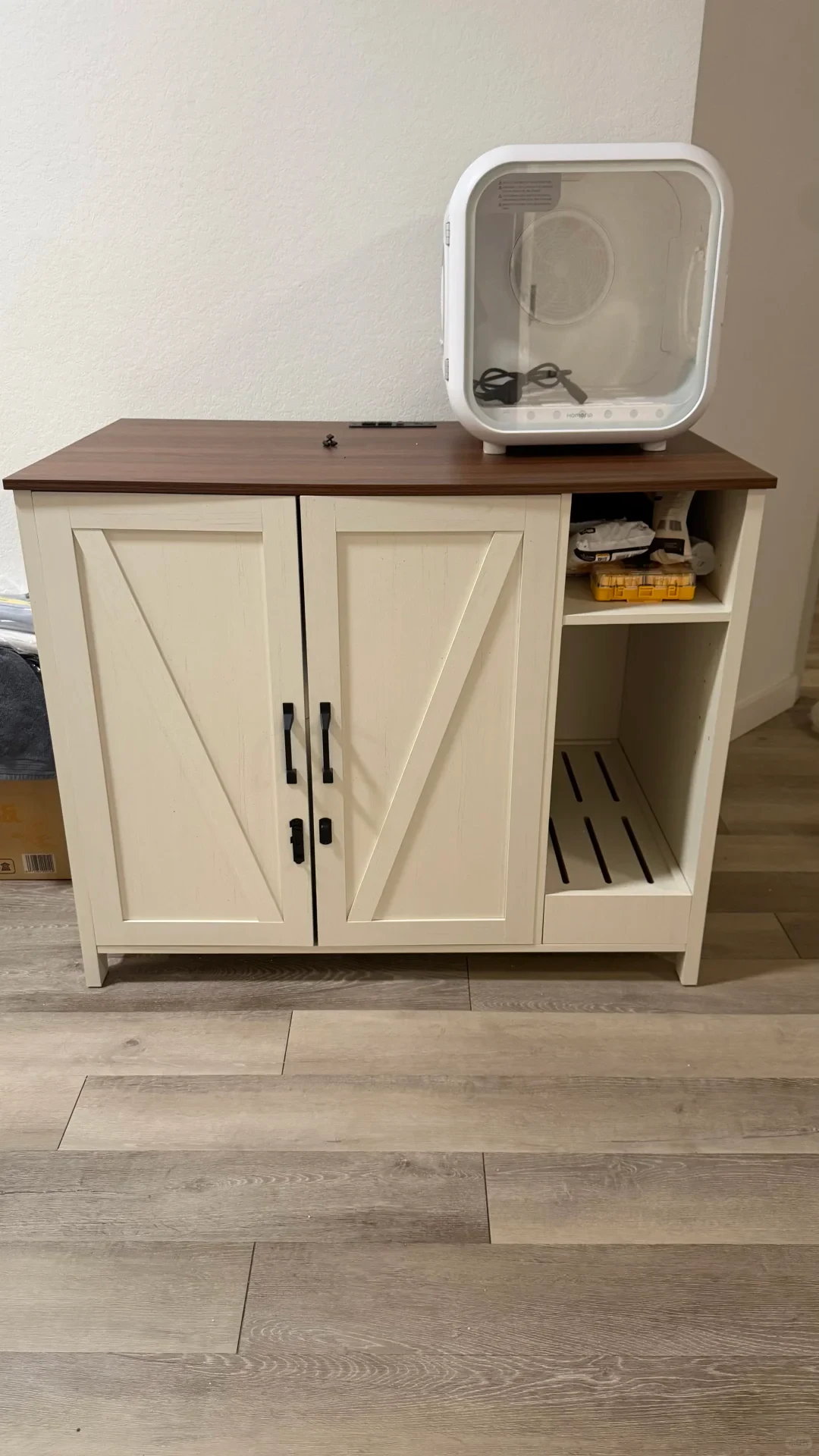 A farmhouse-style cream-colored cabinet with a dark wood top sitting on laminate flooring. The cabinet features double barn-style cabinet doors with black hardware on the left and an open two-tier section on the right. A white smart pet drying box with a clear front door sits atop the right side of the cabinet.