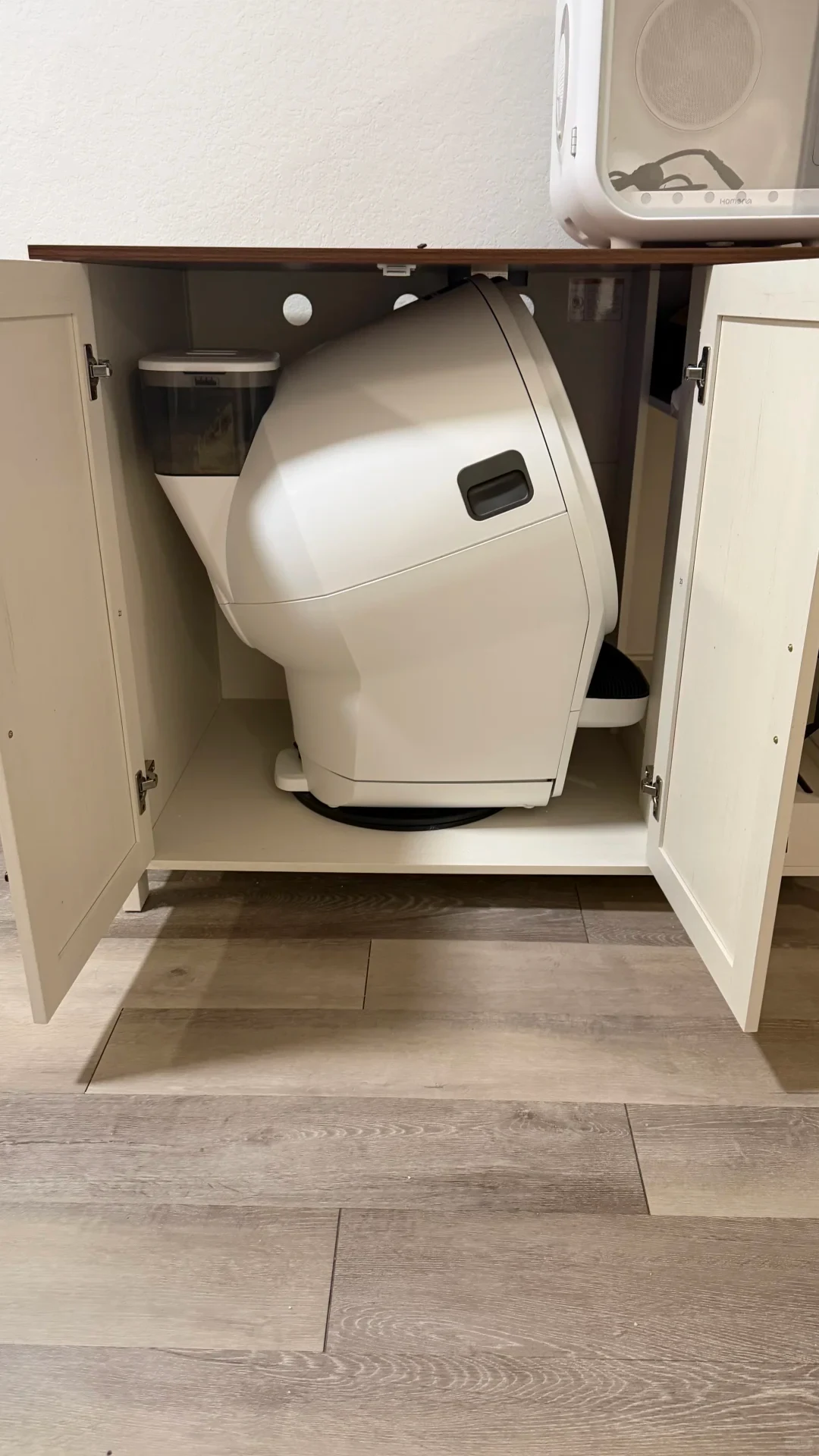 A large white automatic cat litter box wedged at an angle inside a cabinet with its doors wide open, placed over light wood-look laminate flooring.