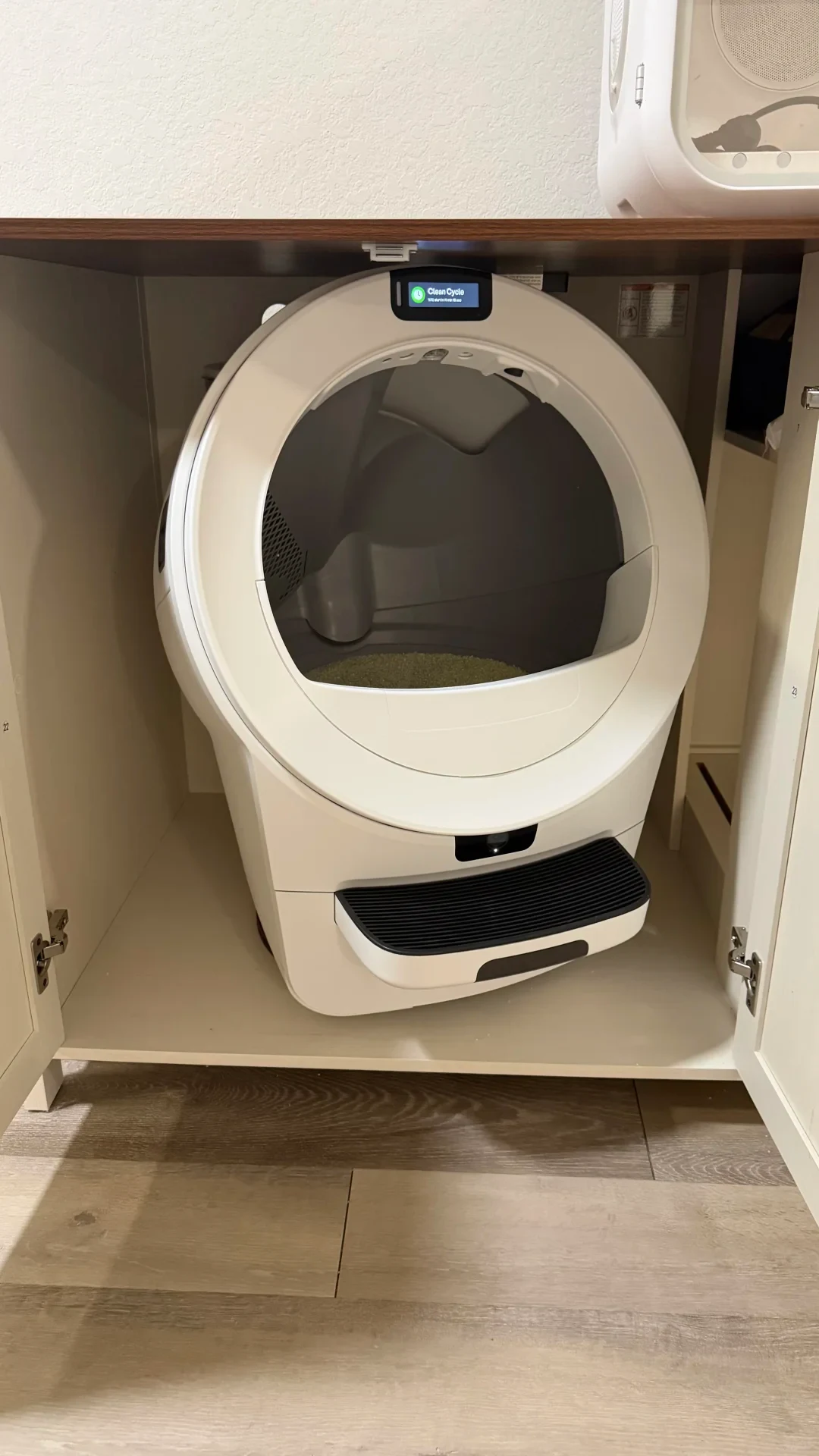 A white automatic cat litter box facing forward inside a cream cabinet with both doors open.