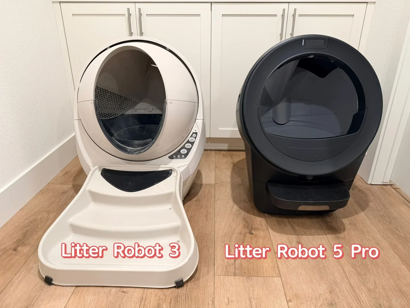 Two automatic litter boxes placed side by side on a light wood floor in front of white cabinets. On the left is a white Litter-Robot unit with attached ramp steps labeled 'Litter Robot 3'. On the right is a sleek, black automatic litter box with a single step labeled 'Litter Robot 5 Pro'.