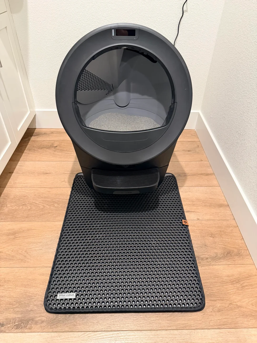 A black automatic cat litter box on a light wood floor, with an entry step and a black honeycomb litter mat in front.