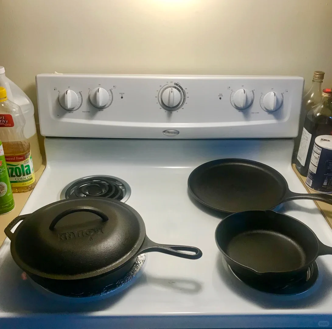 Three black cast iron pans resting on a white electric coil stovetop, including a lidded Lodge skillet on the front left, a small skillet on the front right, and a flat griddle pan on the back right, flanked by bottles of oil and salt.