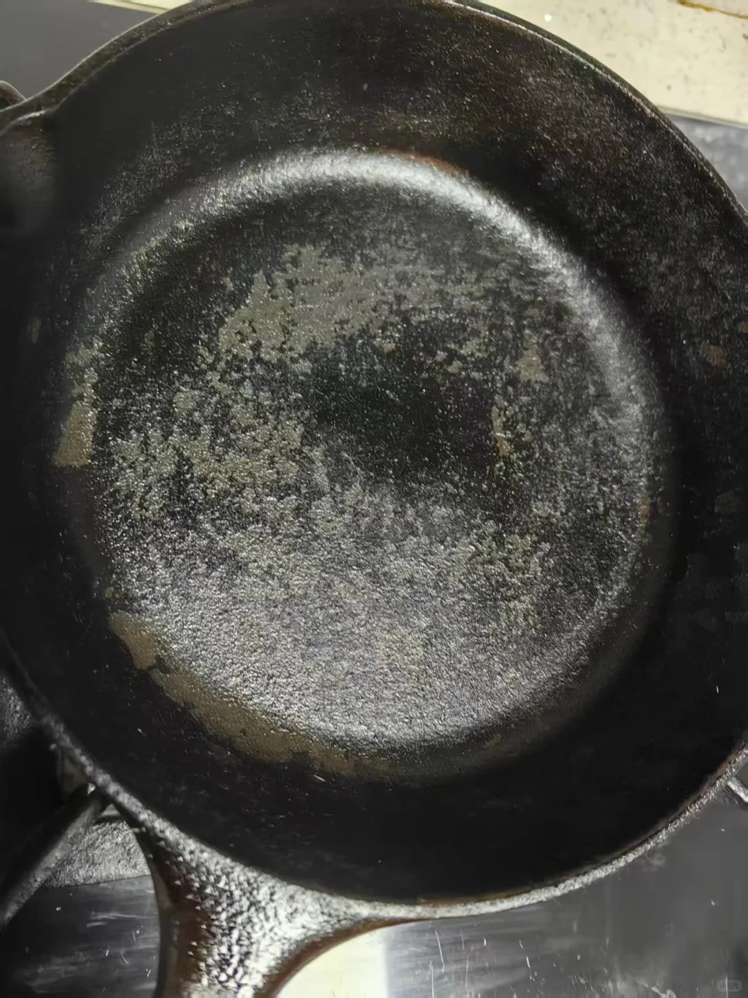 Close-up of a well-used black cast-iron skillet, showing a mottled, worn-looking cooking surface and its handle.