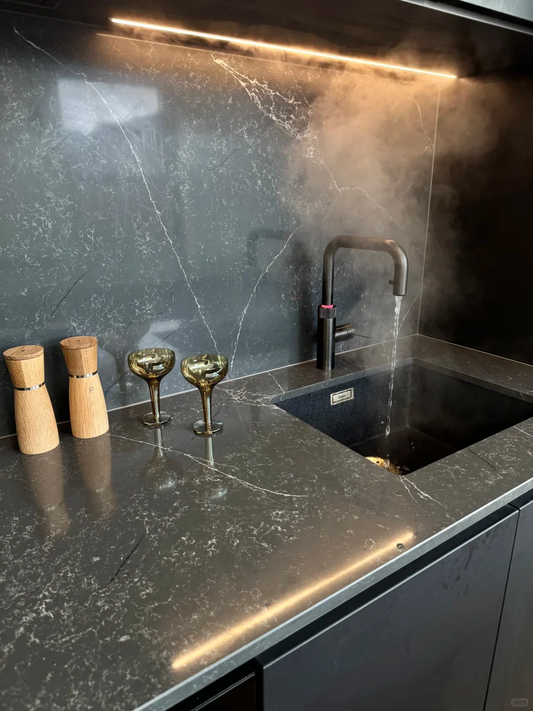 A modern kitchen counter made of dark gray veined stone, where a sleek dark faucet dispenses steaming hot water into an undermount sink under warm under-cabinet strip lighting. Two wooden grinders and two amber coupe glasses sit on the countertop to the left.