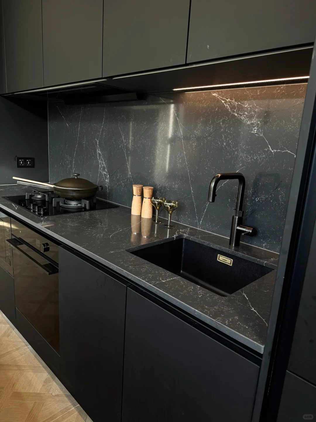 Modern dark kitchen interior featuring black matte cabinetry, a dark marble-patterned quartz countertop and backsplash, an undermount black sink with a matte black faucet, and a built-in cooktop and oven.