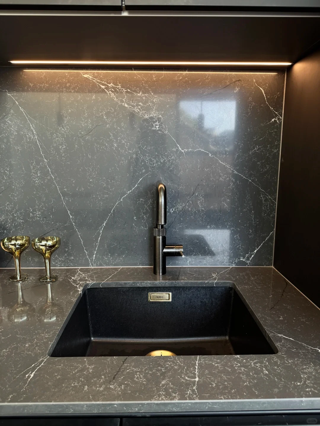A modern undermount black granite composite sink integrated into a dark grey marble countertop and backsplash, featuring a dark brushed metal faucet, under-cabinet lighting, and two golden glasses on the side.