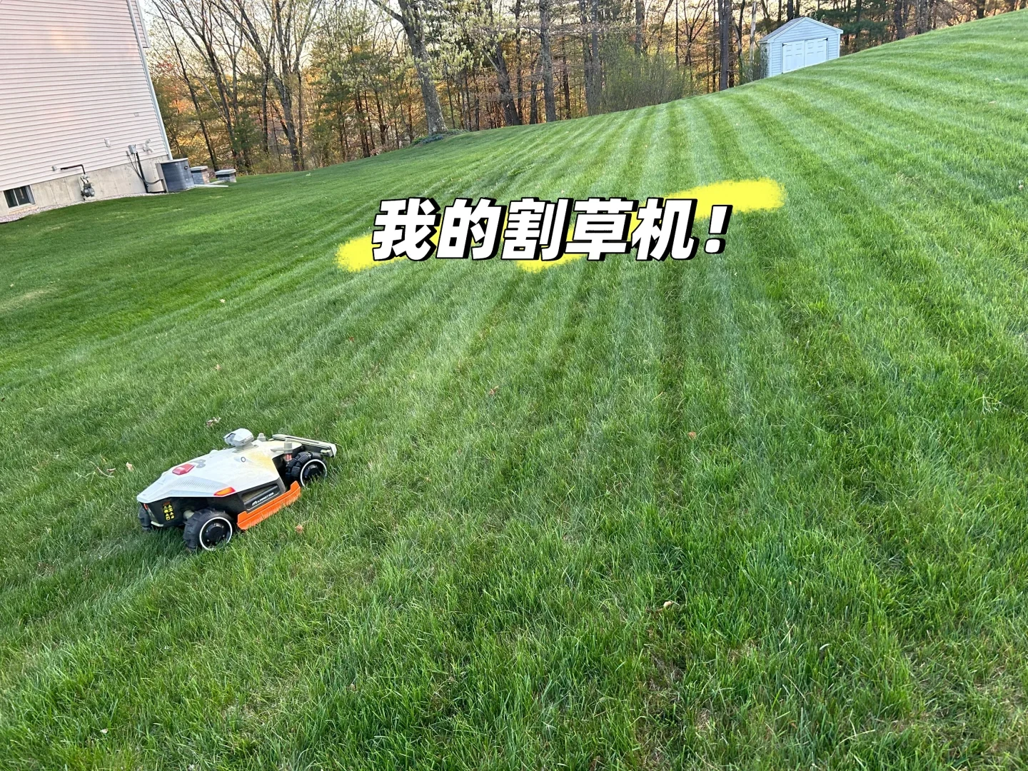 A compact robotic lawn mower operating on a sloped green grass yard, creating neat stripes across the lawn next to a residential house.