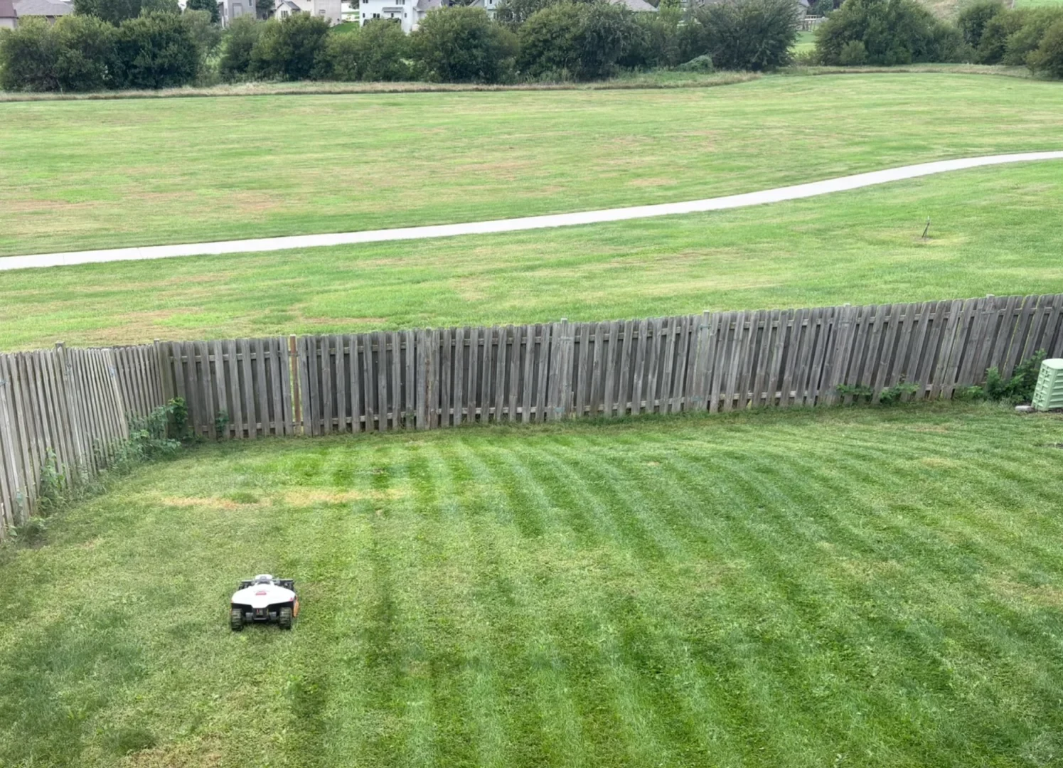 A robotic lawn mower mowing a grassy backyard, creating visible cut striping patterns on the lawn next to a wooden fence, with an open grassy field and paved walking path in the background.