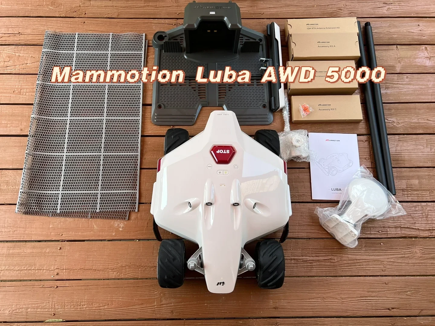 Top-down view of the unboxed Mammotion Luba AWD 5000 robotic lawn mower set arranged on a wooden deck, featuring the white robot mower, charging dock station, mesh floor grid, RTK antenna module, mounting poles, accessory kit boxes, and user manual.
