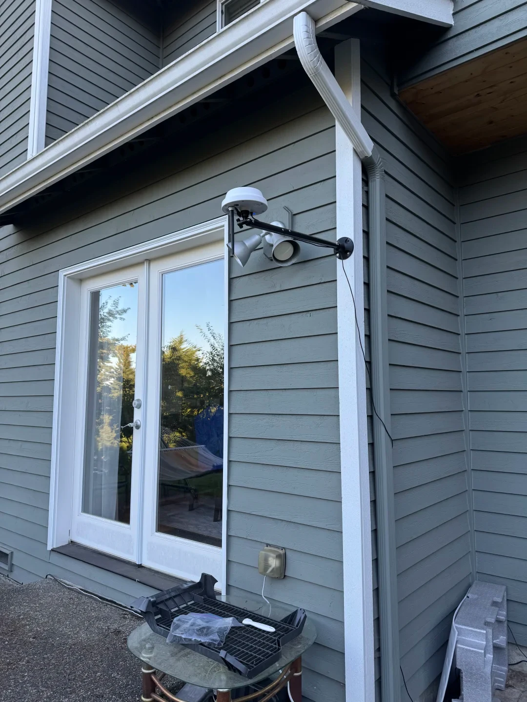 Exterior wall of a gray lap-sided house with white trim around double glass patio doors and a white gutter downspout. Mounted to the wall next to an outdoor floodlight is an adjustable black extension arm holding a small white domed antenna or device, connected via a thin black wire running downward.
