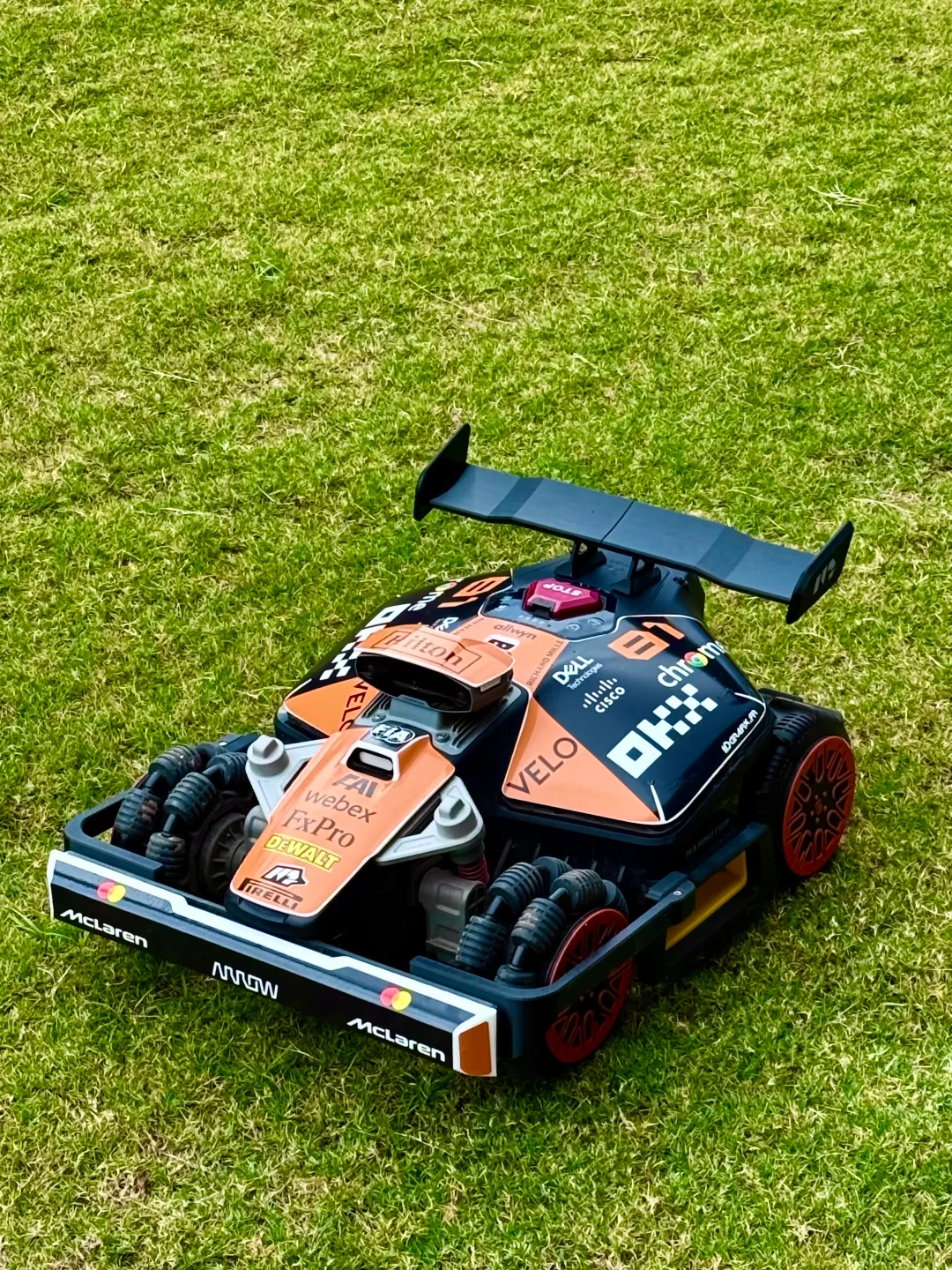 A robotic lawn mower on green grass, custom-designed to resemble a McLaren Formula 1 race car with a papaya orange and black livery, sponsor decals, a rear wing spoiler, and specialized multi-roller wheels.