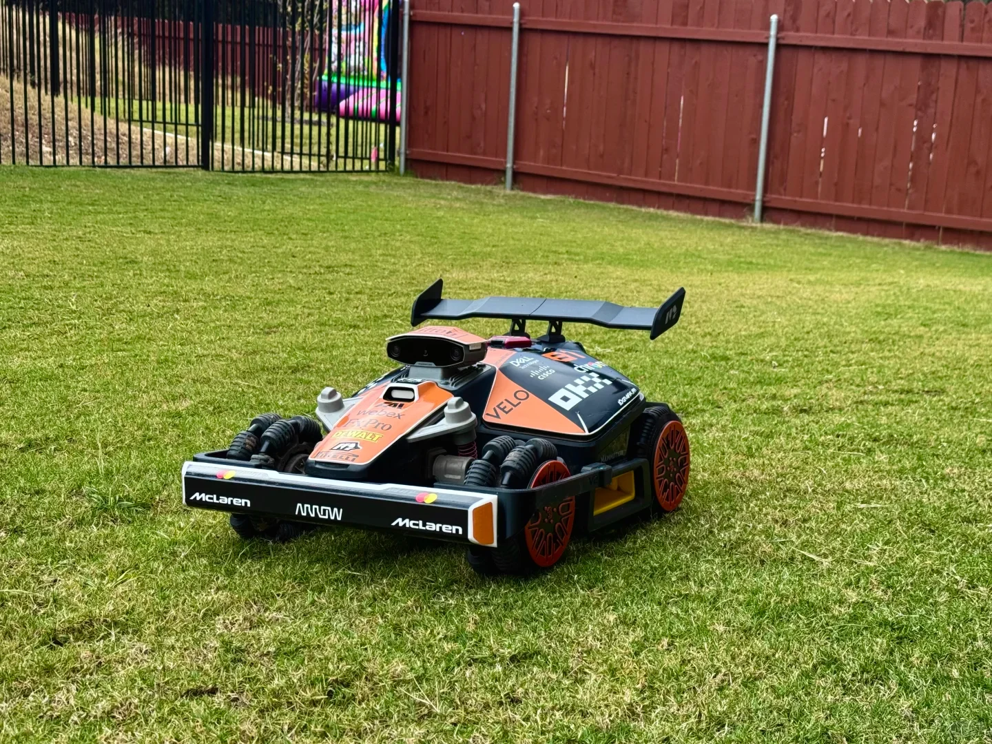 A black and papaya orange robotic rover customized with McLaren Formula 1 race car decals, a rear spoiler wing, and front bumper, resting on a green grass lawn in front of a wooden backyard fence.