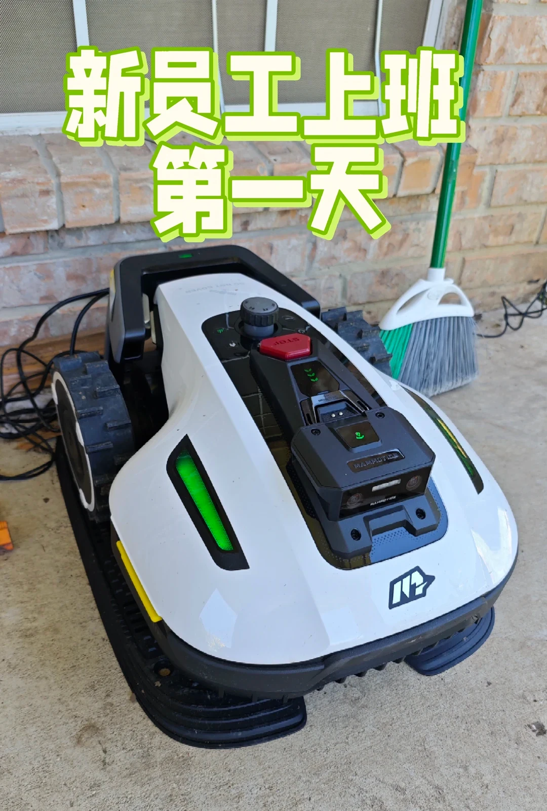 A white Mammotion robotic lawn mower with all-terrain wheels and glowing green LED lights parked on a patio near a brick wall and a broom, beneath large green Chinese text overlay.