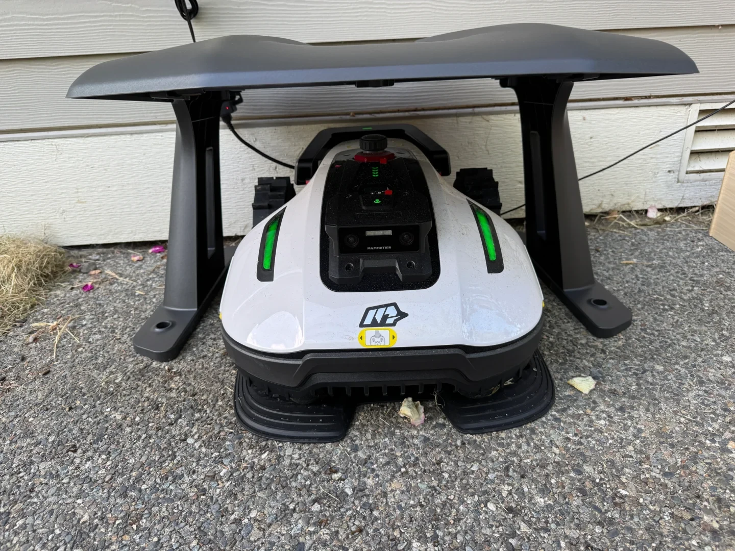 A white and black Mammotion robot lawn mower docked under a black canopy charging station on a paved surface next to house siding.