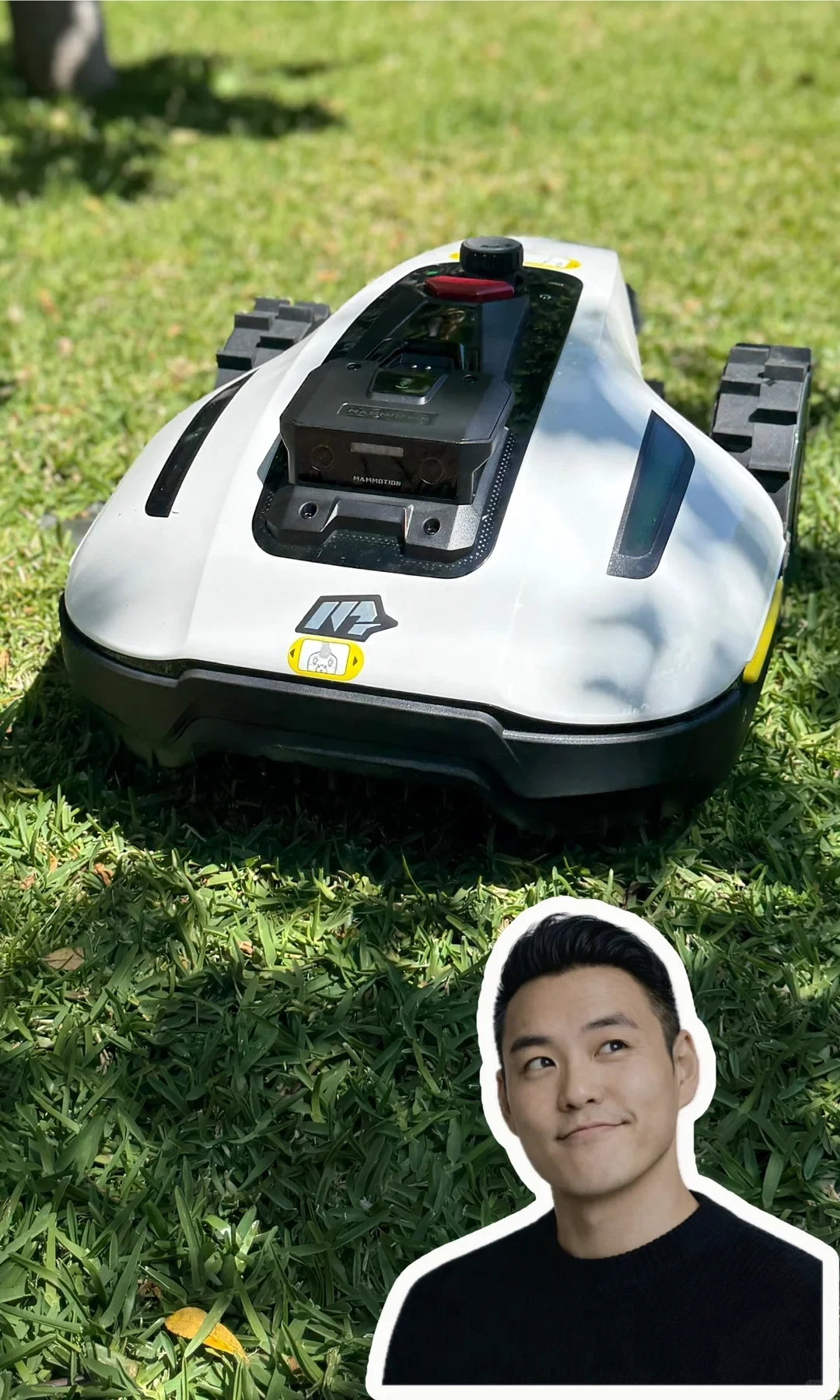 A white Mammotion robotic lawn mower navigating on green grass, with a cutout sticker overlay of a smiling man in the bottom right corner looking up towards it.