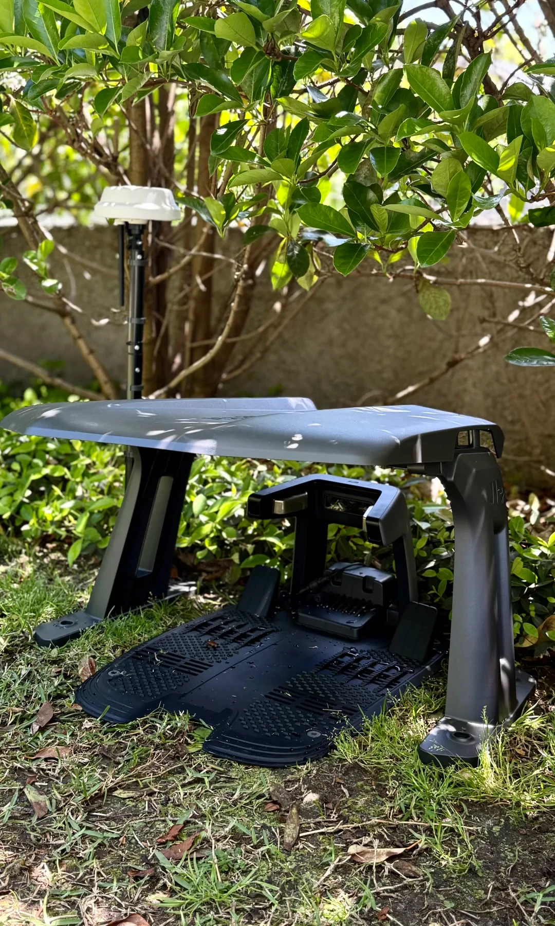 A dark gray robotic lawn mower base station sheltered beneath a matching plastic canopy on a lawn, next to an RTK reference antenna mounted on a pole near garden bushes.