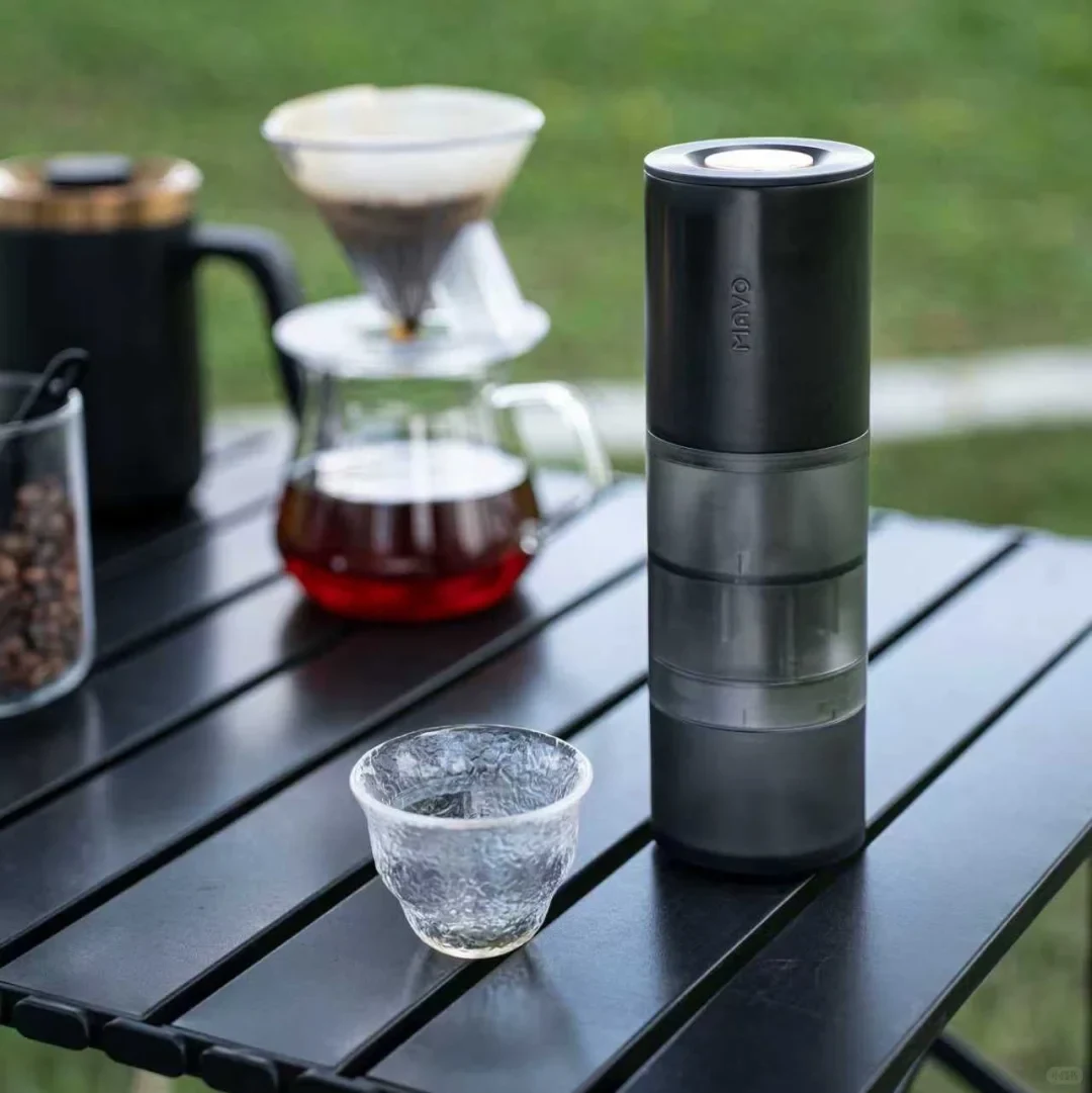 Product-context photo of a black MAVO Aurora grinder on an outdoor slatted table beside a textured glass cup, with a pour-over dripper and server behind it.