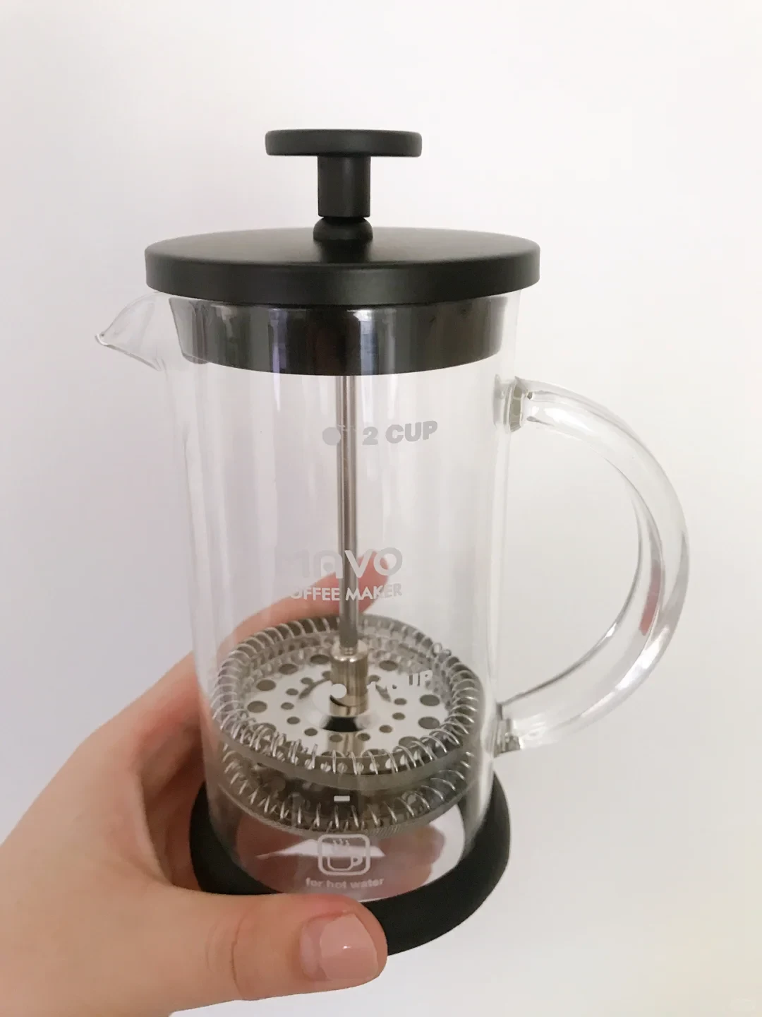 A hand holds a clear, two-cup French press coffee maker with a black lid and base, glass handle, and visible metal plunger filter.