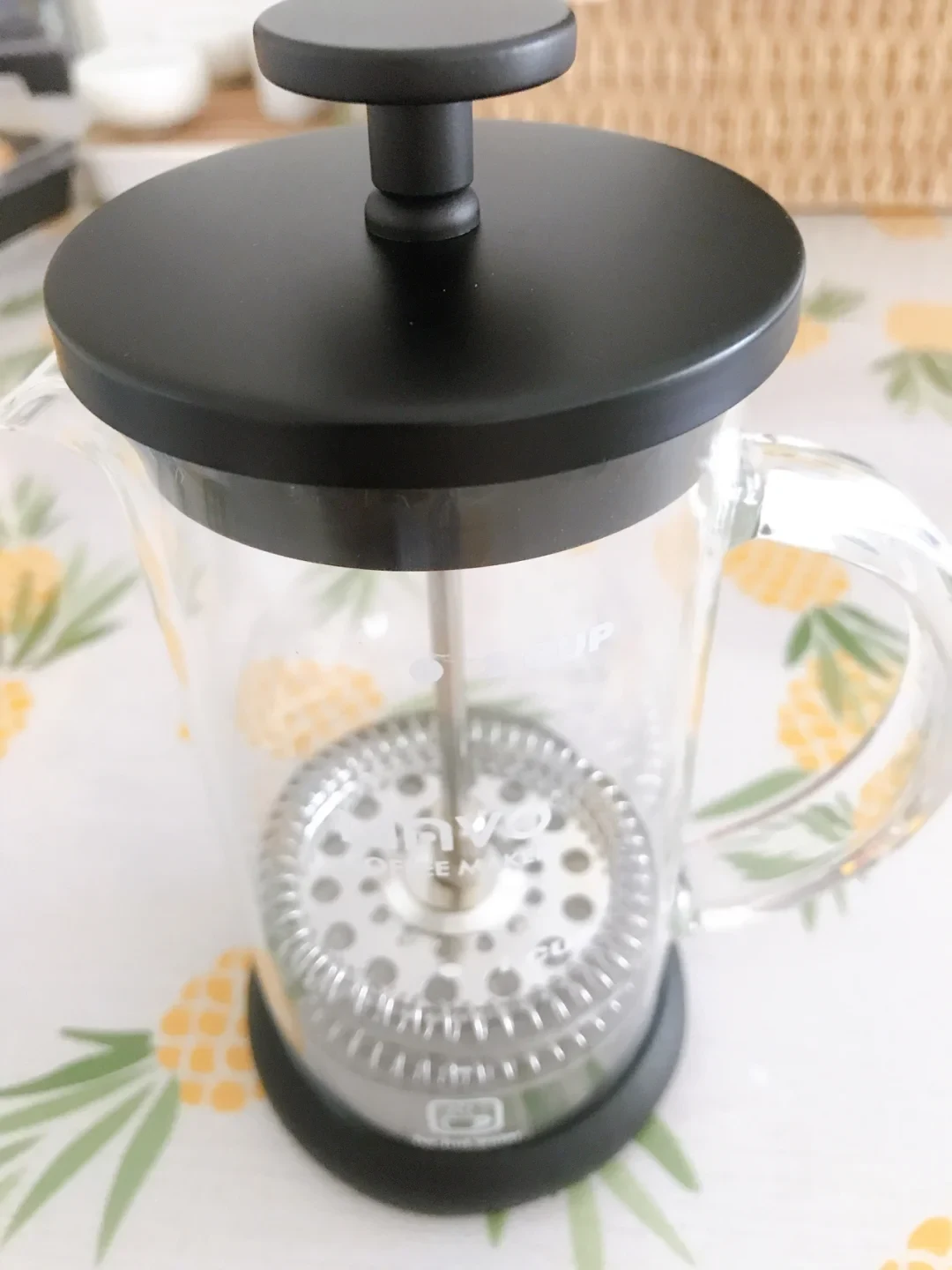 A clear glass French press with a black lid and base, a plunger rod, and a perforated metal filter sits on a fruit-patterned tablecloth.