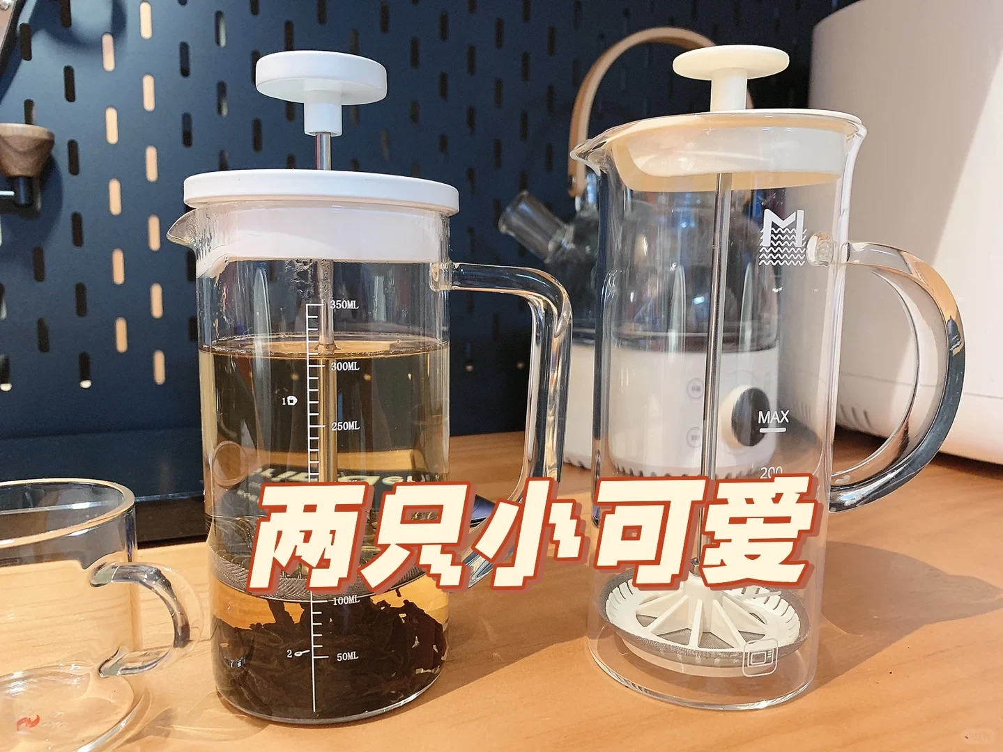 The owner’s Coffee Plus French press, holding tea on the left, stands beside a MAVO Snow Meet milk foamer on the right. Both have clear glass bodies and light-colored lids; the source overlay calls them two little cuties.