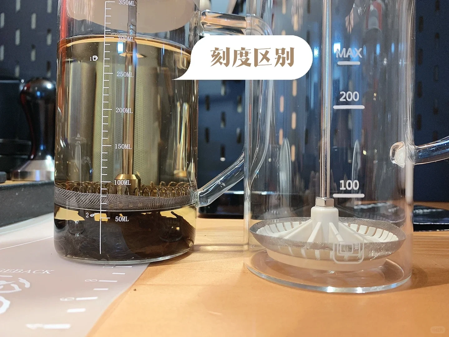 A product comparison photo shows two clear glass press pots on a wooden counter: the left pot contains dark liquid and has a fine milliliter scale, while the empty pot on the right has larger 100, 200, and MAX markings. A speech-bubble label highlights the difference in their graduations.