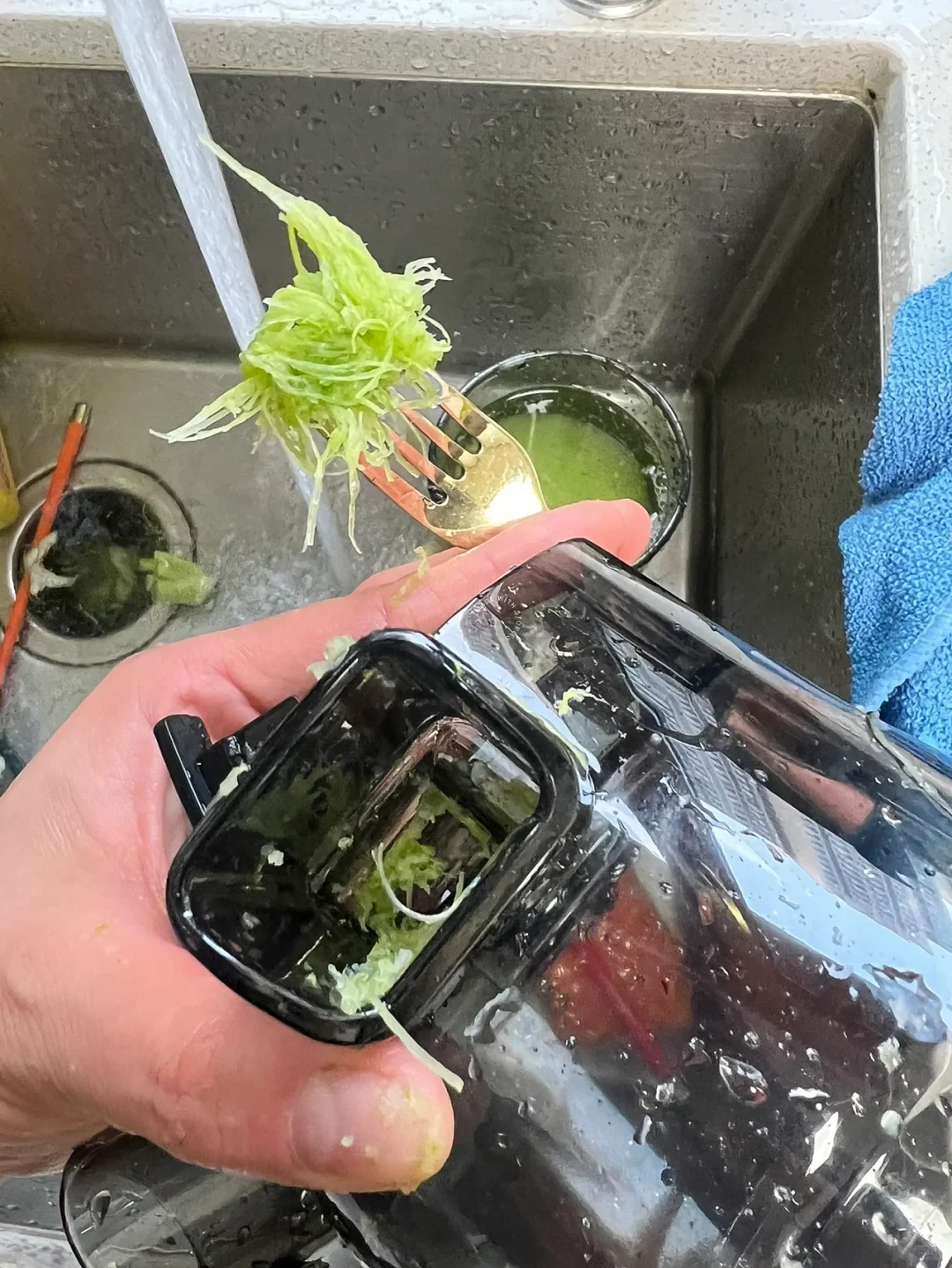 A hand holds a wet black-and-clear kitchen appliance part over a sink while shredded green vegetables are rinsed under running water.