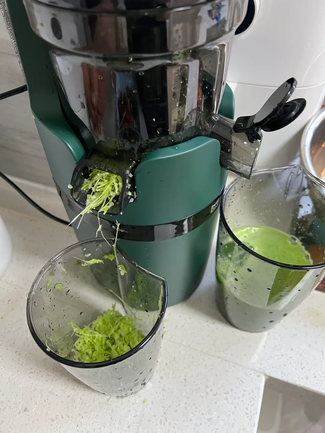 A dark green juicer is extracting leafy greens, with shredded pulp dropping into a clear cup and green juice collecting in another cup beside it.