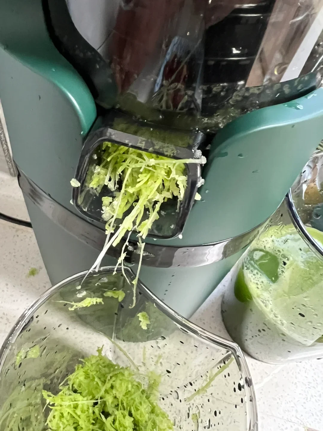 A teal juicer is processing leafy green vegetables, with shredded pulp emerging from its front outlet into a clear container and green juice collecting in a cup beside it.