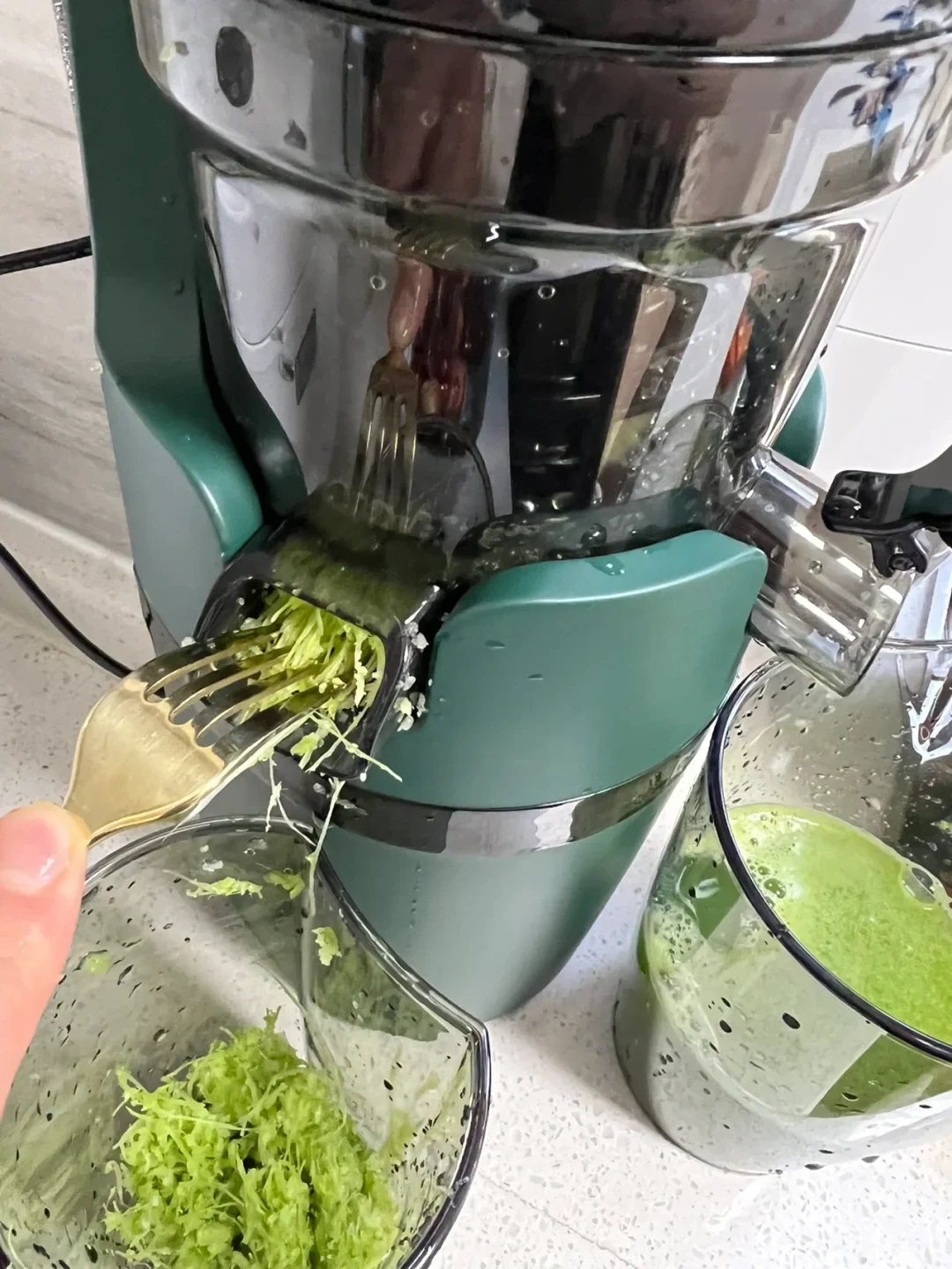 A teal-and-stainless-steel juicer is processing leafy green vegetables; shredded pulp exits into a clear container while green juice collects in a separate cup.