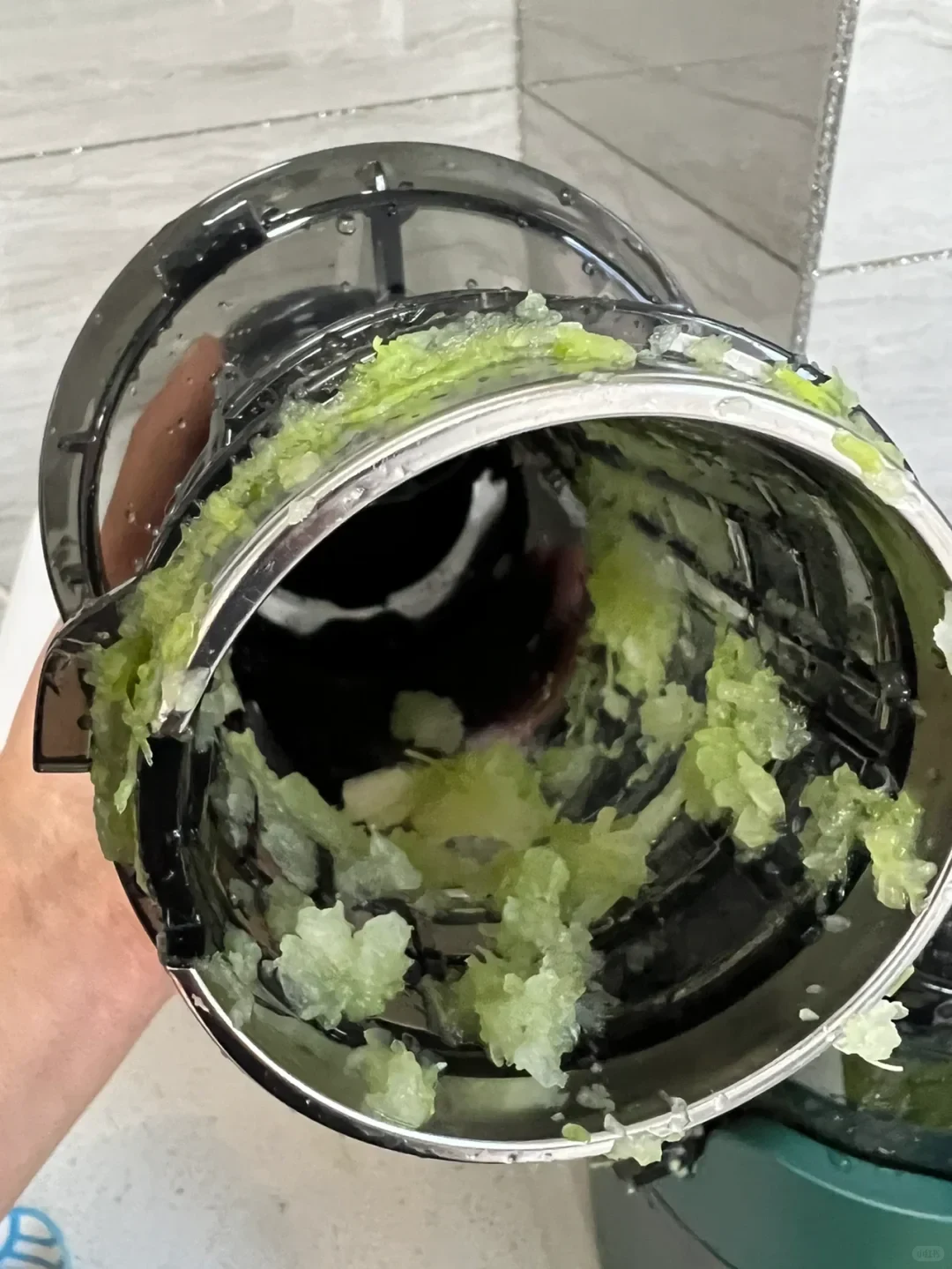 A hand holds open a circular appliance component coated inside with clumps of pale green residue; a tiled wall and part of the appliance are visible behind it.
