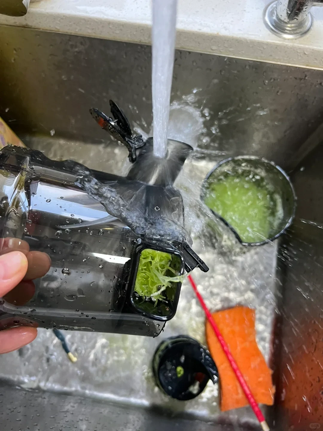 A hand holds a black-and-clear kitchen device under running water in a sink, rinsing green pulp from its open compartment. A strainer with green residue and an orange sponge are nearby.
