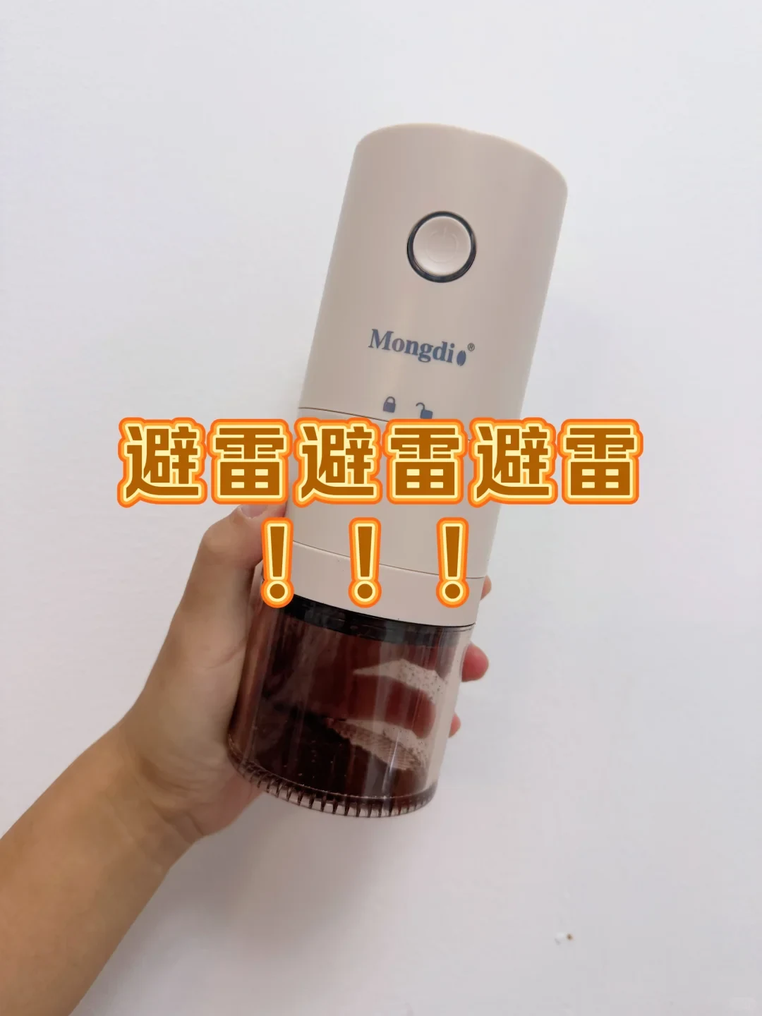 A hand holds a cream Mongdio portable electric coffee grinder with a round button and a transparent grounds jar. The source’s Chinese overlay repeats a warning to avoid it.