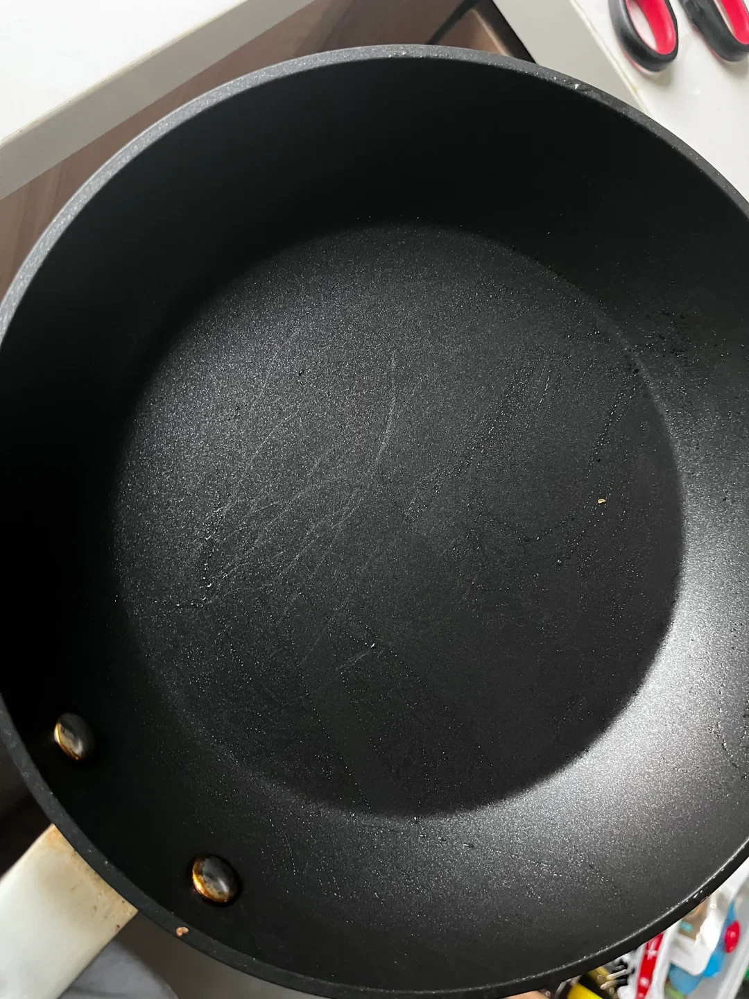 Top-down view of the inside surface of a black non-stick frying pan, showing faint scratches on the bottom and two rivets connecting the handle at the lower left.