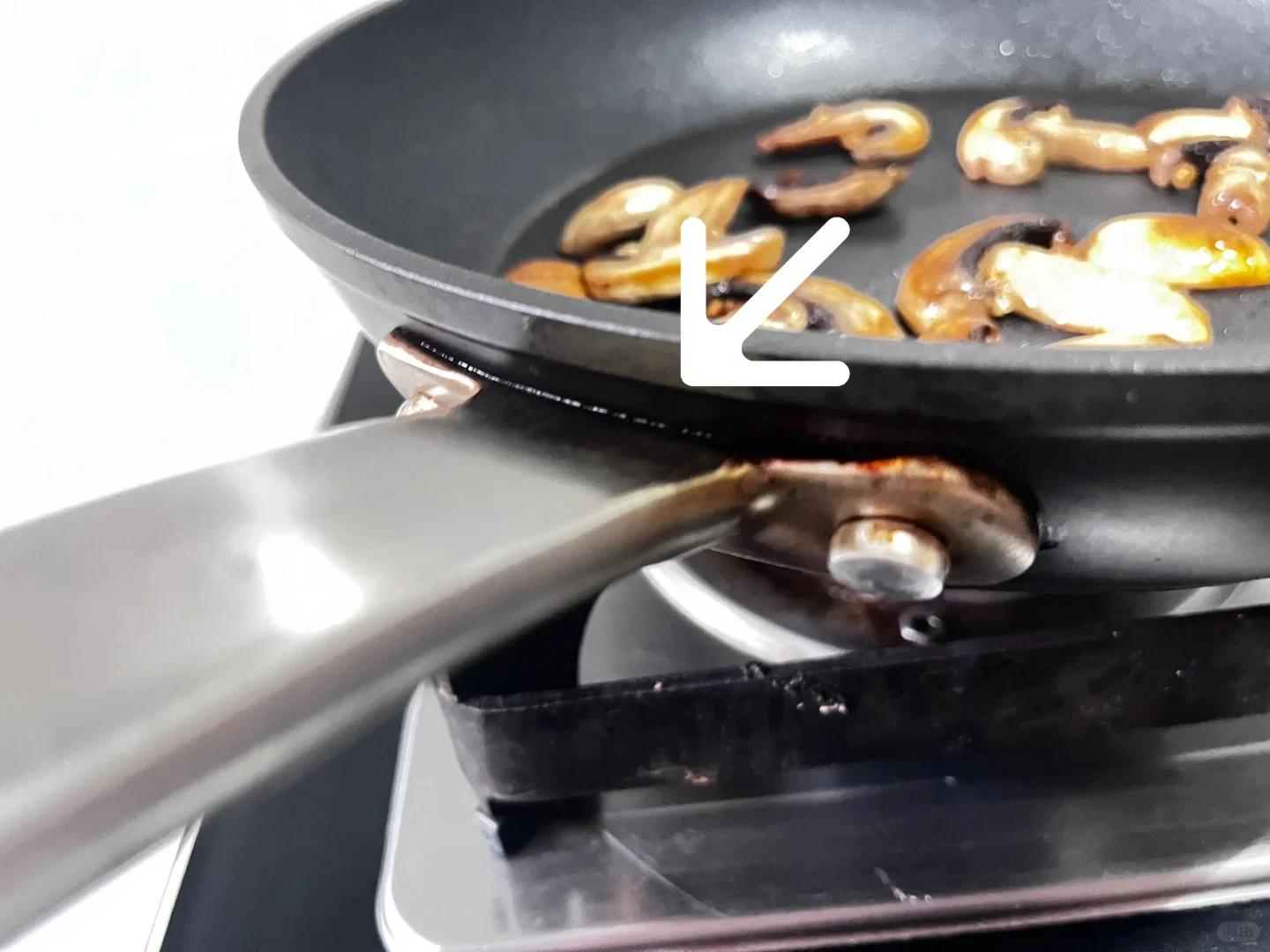 A frying pan with sliced mushrooms cooking inside, featuring a white arrow pointing to scorching and grease buildup at the metal handle attachment.