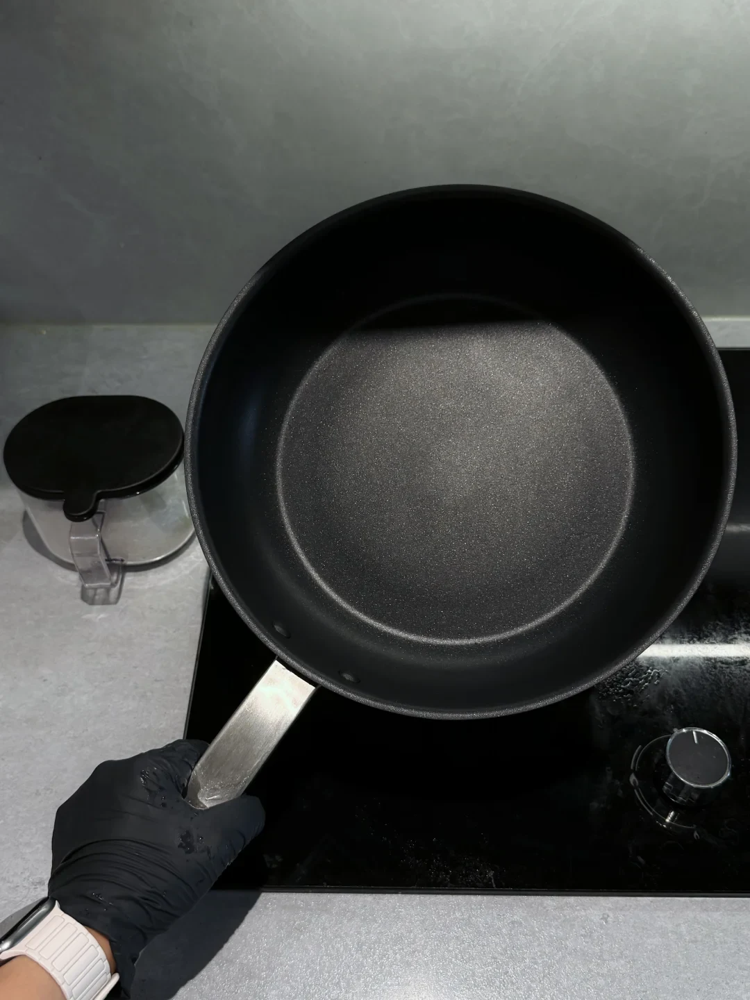 A black nonstick frying pan is held upright by its metal handle over a black glass cooktop; a small covered container sits on the counter to the left.
