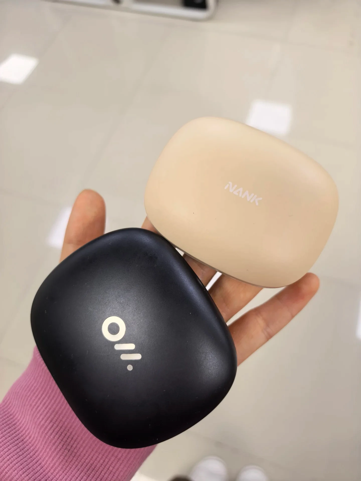 A hand holding two wireless earphone charging cases side by side, one in matte black with a silver logo, and one in beige printed with the brand name NANK.