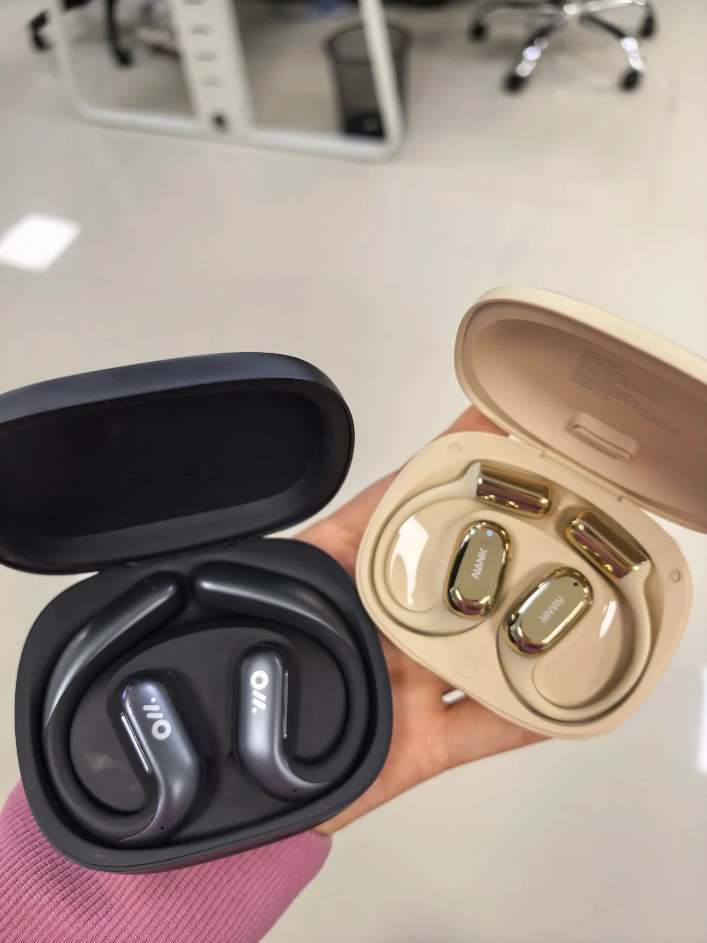 A person holds two open charging cases for open-ear wireless earphones: a black case on the left containing dark metallic earhook earbuds, and a beige case on the right containing gold-accented earbuds branded NANK.