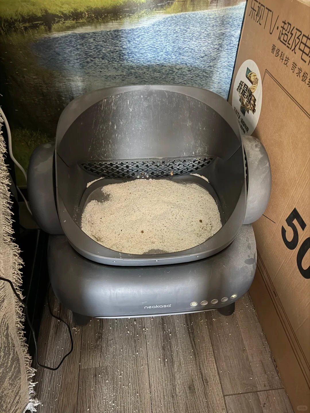 A dark gray Neakasa automatic cat litter box filled with litter, placed on a wood floor next to a cardboard TV box.