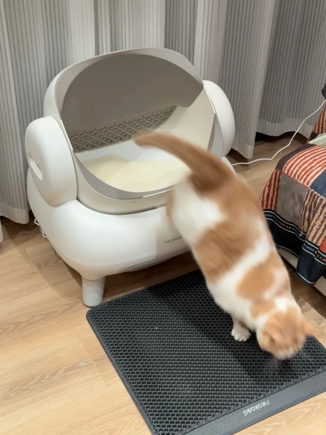 An orange and white cat jumps out of an open-top, automatic self-cleaning litter box onto a black litter trapper mat on a wooden floor.