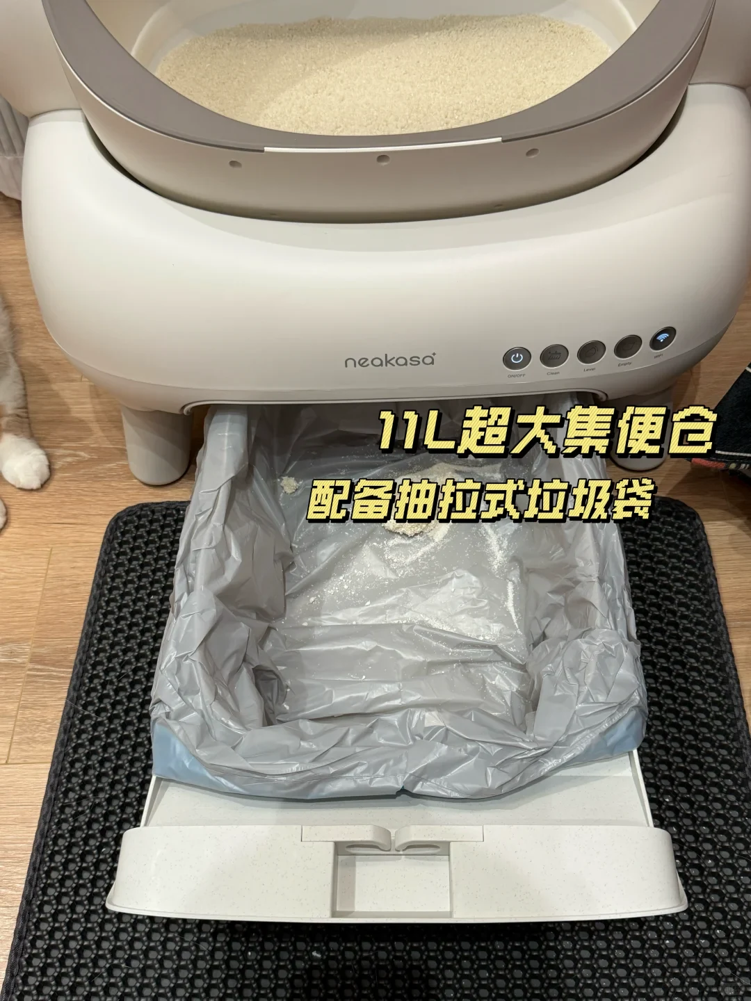 A white Neakasa automatic cat litter box with its lower waste compartment pulled out onto a black honeycomb litter mat, showing a gray plastic trash bag lining the drawer.