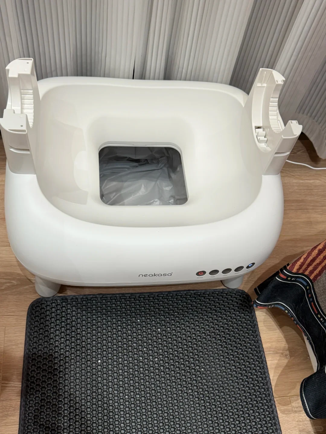 Top view of a disassembled white Neakasa automatic cat litter box base resting on a wood floor, featuring a central opening lined with a waste bag, mounting brackets on both sides, control buttons on the front rim, and a black honeycomb litter-trapping mat placed in front.