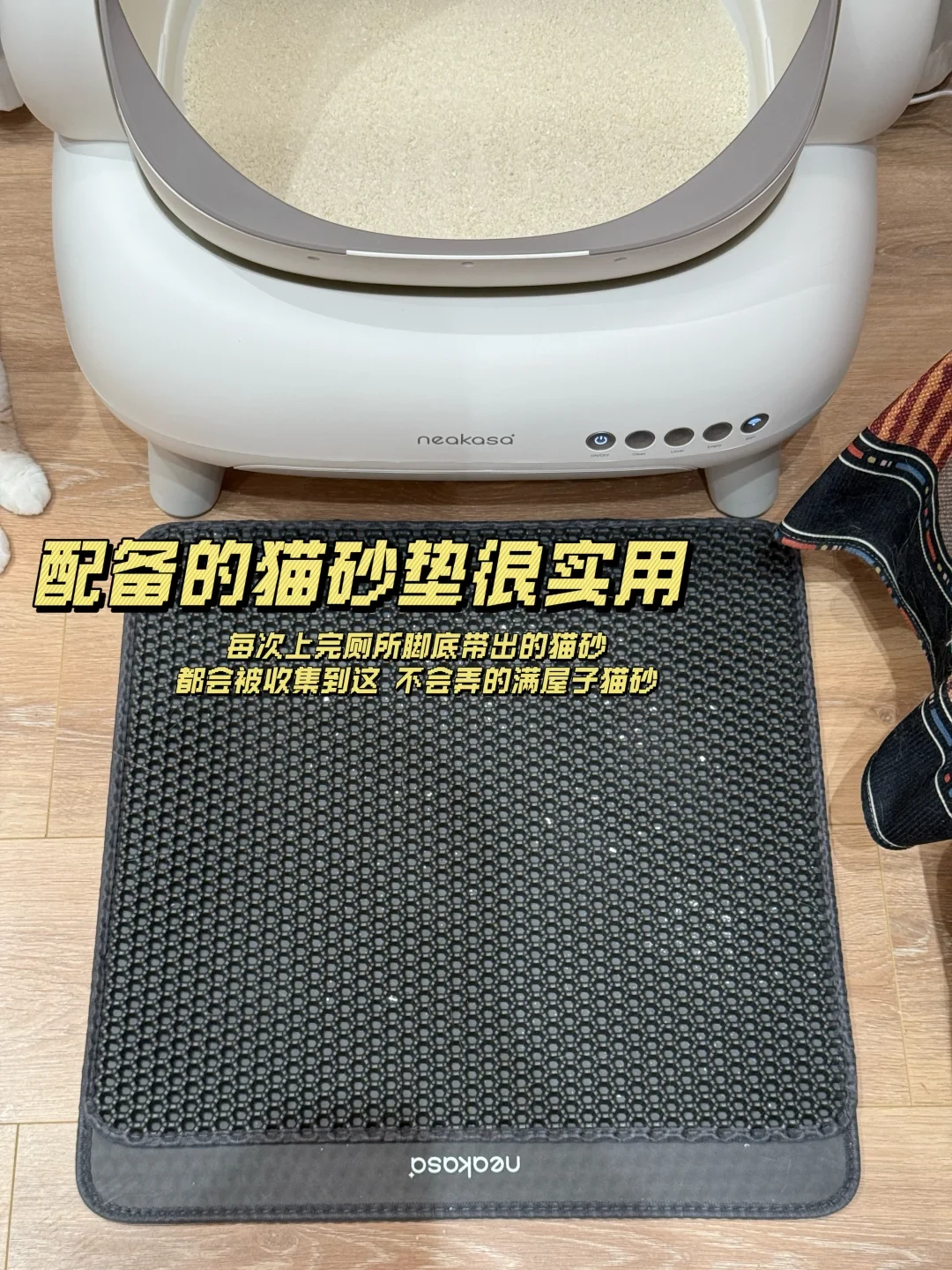 A top-down view of a white Neakasa smart cat litter box on a light wood floor, with a dark gray honeycomb cat litter trapping mat placed directly in front of the entrance.