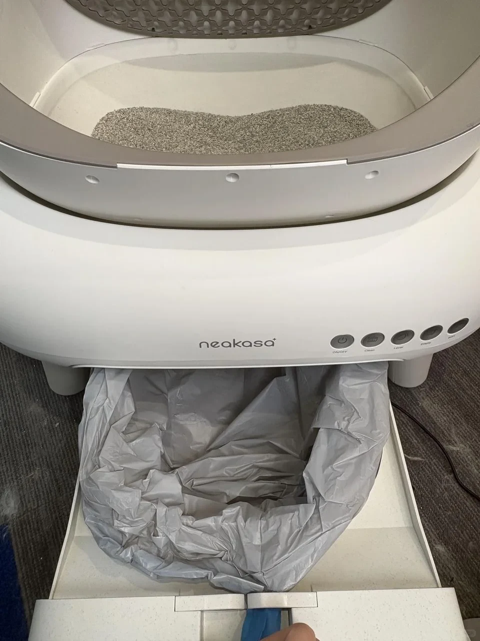 A white Neakasa automatic litter box with its bottom waste compartment drawer pulled out, fitted with a gray plastic liner bag below the open litter chamber.