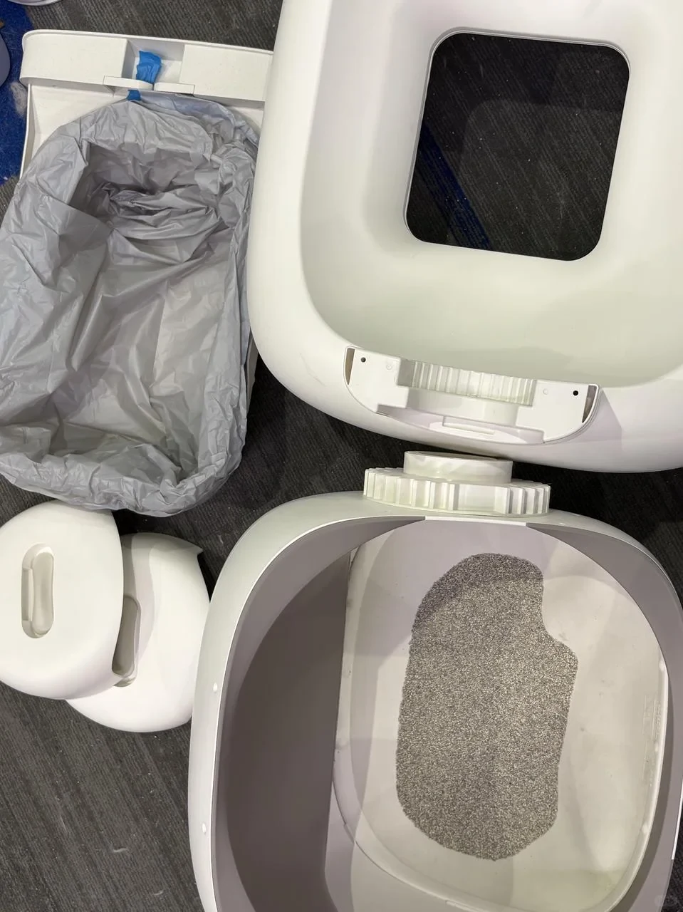 Disassembled white components of an automatic cat litter box laid out on a dark carpet, showing the inner litter drum with cat litter inside, the upper frame with gear mechanism, a waste bin lined with a gray trash bag, and side covers.