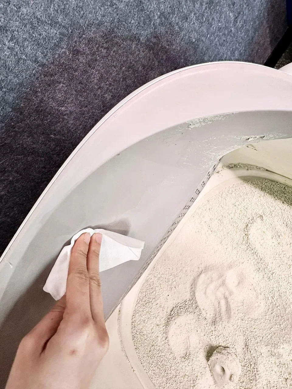 A close-up of a person's hand using a white wet wipe to clean the inner curved rim of an automatic cat litter box filled with litter.