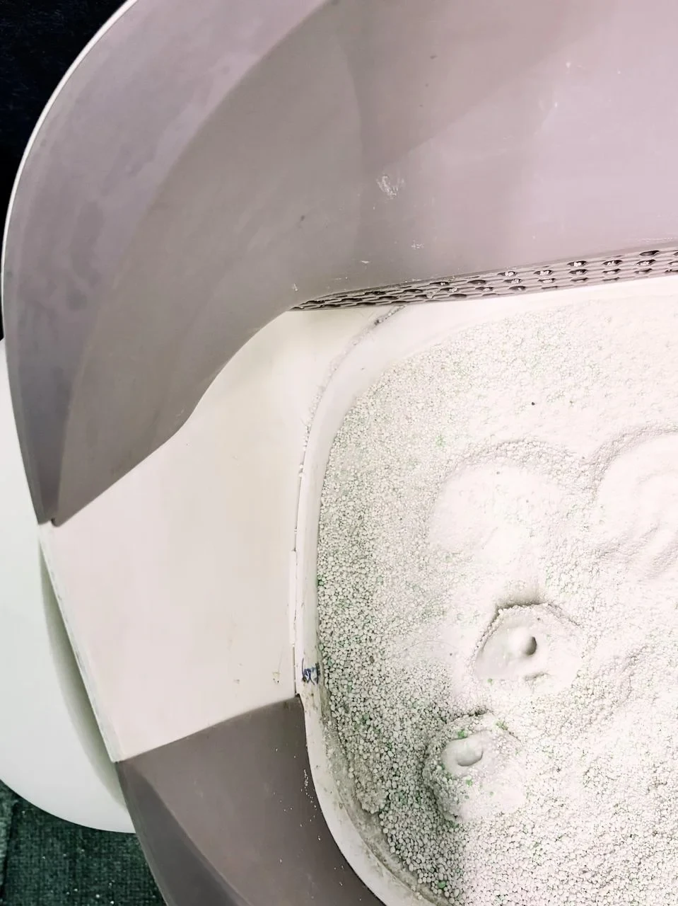 Top-down close-up view into the open drum of an automatic cat litter box filled with fine white and green litter with cat paw impressions next to the internal sifting grate.