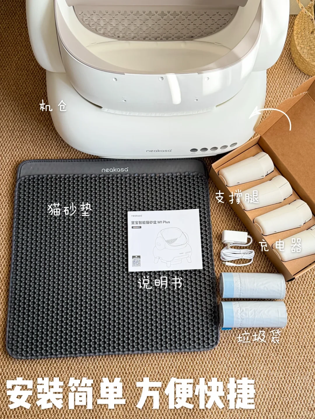 A Neakasa M1 Plus smart cat litter box unboxed and displayed with accessories on a woven rug. The white litter basin sits at the top, accompanied below by a dark gray litter-trapping mat, an instruction booklet, a cardboard box containing four white support legs, a white power charger, and two rolls of gray waste bags. White Chinese annotations label each component and provide a promotional caption at the bottom.