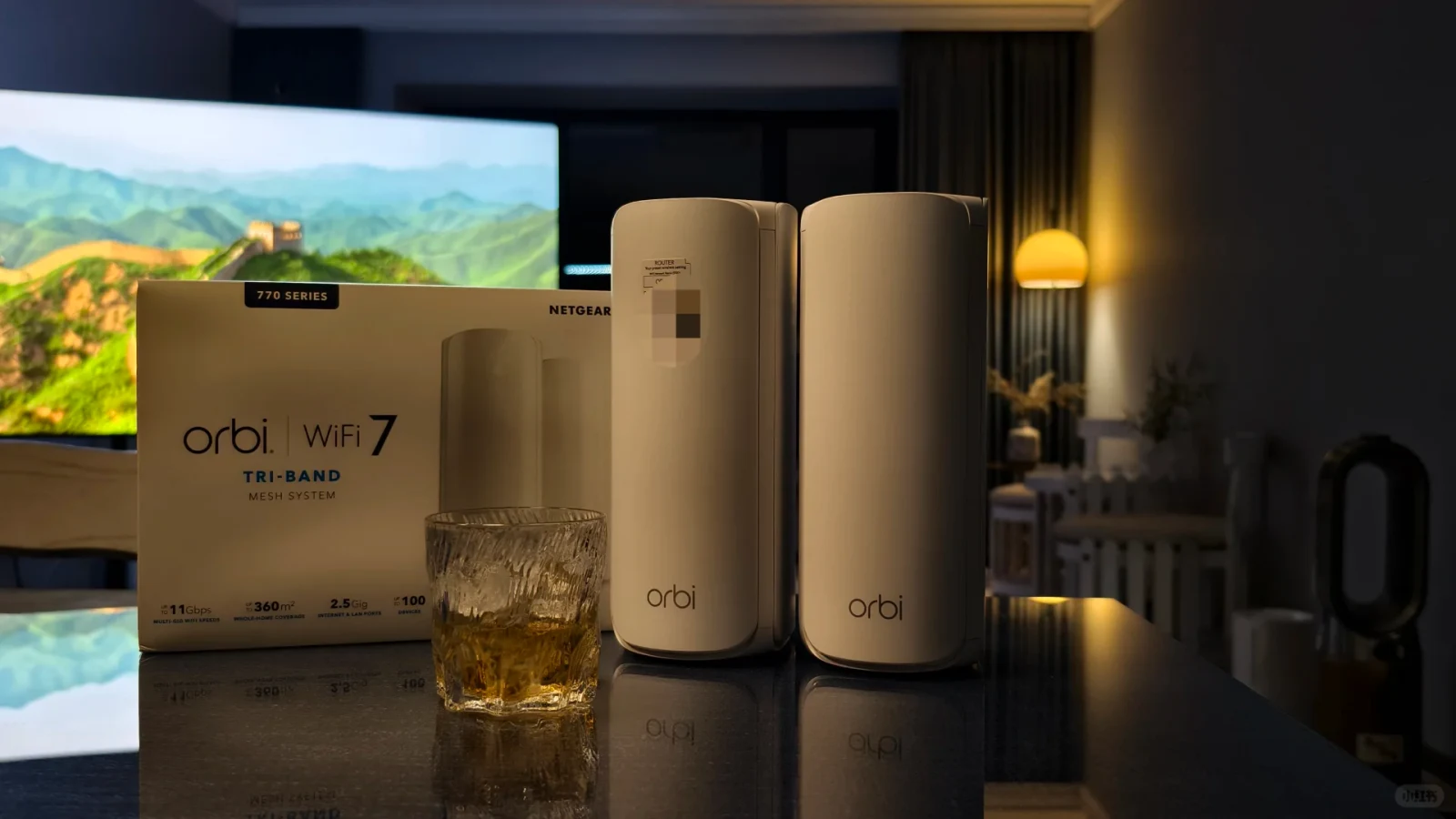 Two white Netgear Orbi 770 Series mesh router towers placed next to their product box and a glass of iced drink on a glossy dark table.