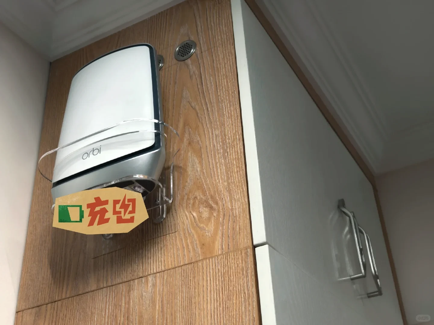 A white Netgear Orbi mesh Wi-Fi router mounted high on a wood-paneled cabinet in a clear plastic wall mount, featuring a decorative sticker on the lower left.
